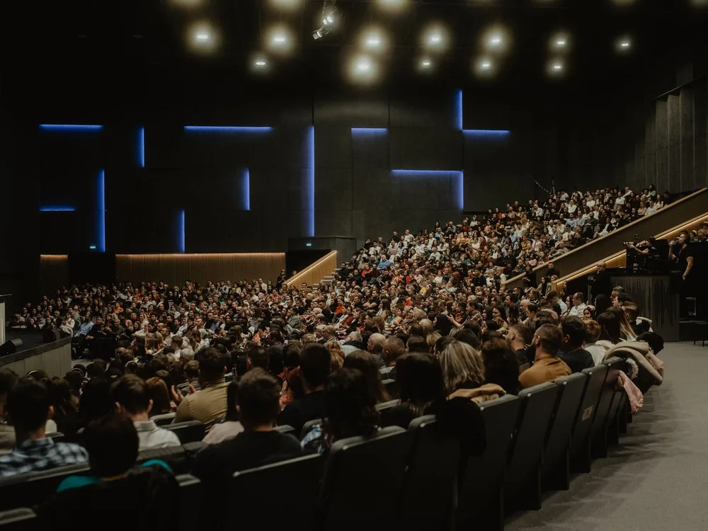 Auditorio corporativo en penumbra con la platea completa y un orador en el escenario